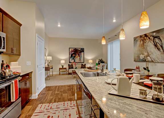 A welcoming kitchen here at 4800 Excelsior featuring an elongated island with light stone countertops, an integrated sink, and modern place settings beneath warm pendant lighting. Dark wood cabinetry and stainless steel appliances frame the space, with wood-style flooring flowing into an open living area accented by seating, artwork, and large windows.