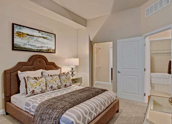 A serene bedroom here at 4800 Excelsior featuring a plush upholstered bed with patterned bedding, layered accent pillows, and a textured throw. Soft carpeting and neutral walls create a calming atmosphere, complemented by a bedside lamp, framed artwork, and a mirrored nightstand, with easy access to a walk-in closet and adjoining bathroom.
