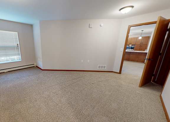 A spacious, empty living room here at Club at Heritage with soft carpet and neutral walls. A large window with blinds brings in natural light, and an open doorway leads into the kitchen for a connected, functional layout.