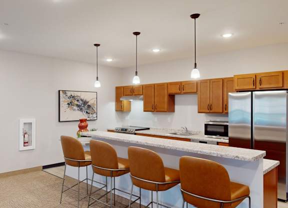 A modern clubhouse kitchen here at Club at Heritage with wood cabinetry, stainless steel appliances, and granite-style countertops. A large breakfast bar with seating and pendant lighting creates a stylish, functional space for gathering.
