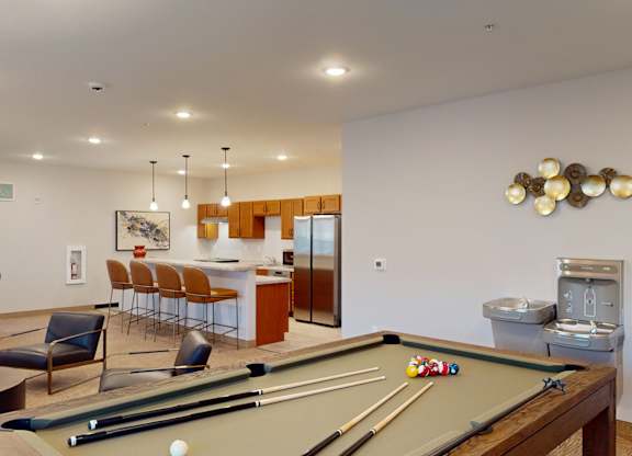 A community clubhouse here at Club at Heritage with a pool table, lounge seating, and a modern kitchen area. Pendant lighting, bar seating, and open space create a fun and inviting environment for gathering and entertainment.