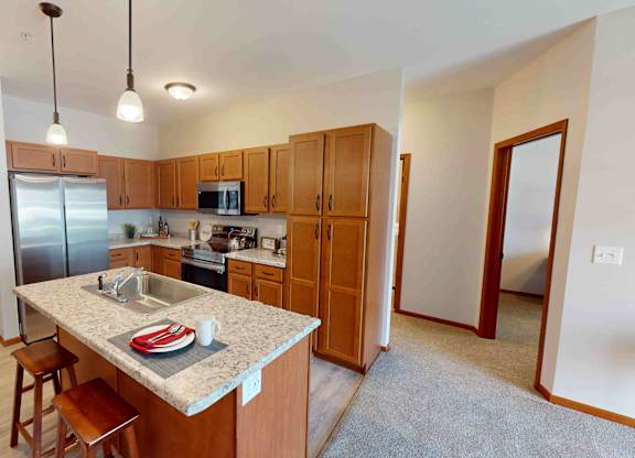 A kitchen here at Club at Heritage featuring warm wood cabinetry, stainless steel appliances, and a spacious island with a built-in sink and seating, opening into a bright, carpeted living area.