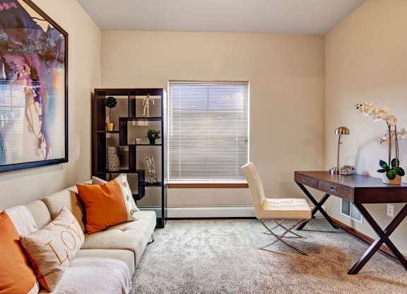 A home office here at Club at Heritage featuring a cozy workspace with a wooden desk, upholstered chair, decorative shelving, and a soft neutral sofa with orange accent pillows.