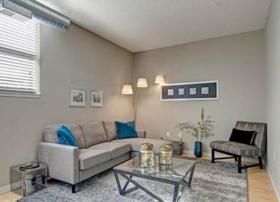 A cozy living room here at Marbella on Dean featuring a light gray sofa with blue accent pillows, a glass coffee table centered on a soft area rug, and neutral walls complemented by framed artwork, with an upholstered accent chair, modern floor lamp, exposed ductwork, and a window with blinds bringing in natural light.