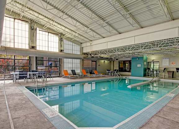 A spacious indoor pool here at Marbella on Dean featuring a large rectangular swimming pool with clear blue water, stainless steel handrails, surrounding concrete deck, and lounge seating with bright accent chairs, set beneath a high industrial-style ceiling with exposed beams and large windows that bring in natural light.