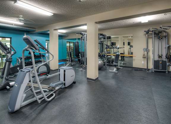 A spacious fitness center here at Marbella on Dean featuring cardio machines and strength equipment arranged across dark rubber flooring, mirrored walls, and teal accent walls, with weight racks, functional training space, and bright overhead lighting creating an open and motivating environment for daily workouts.