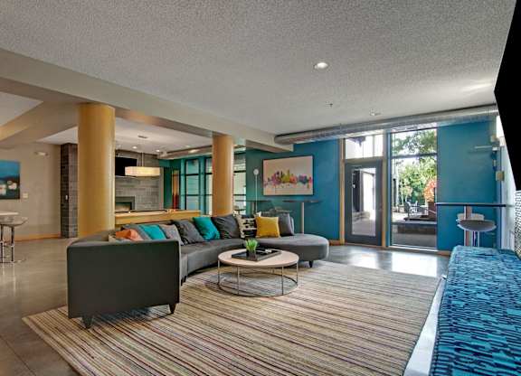 A modern community lounge here at Marbella on Dean featuring a large sectional sofa with colorful accent pillows arranged on a striped area rug, round coffee tables, and warm wood columns, with teal accent walls, contemporary artwork, high-top seating, and floor-to-ceiling windows and glass doors that bring in natural light and connect to an outdoor patio.
