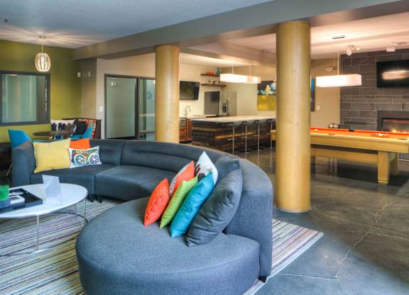 A stylish community lounge here at Marbella on Dean showcasing a curved sectional sofa with vibrant accent pillows arranged on a striped area rug, paired with round coffee tables, warm wood columns, and polished concrete floors, with a billiards table, modern fireplace, bar-style seating, and soft ambient lighting creating an inviting social space.