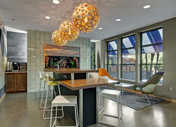A stylish community gathering space here at Marbella on Dean featuring a long wood communal table with modern bar seating, sculptural pendant lighting overhead, and polished concrete flooring, complemented by a built-in aquarium wall, large windows with natural light, and cozy lounge chairs that create a social and inviting atmosphere.