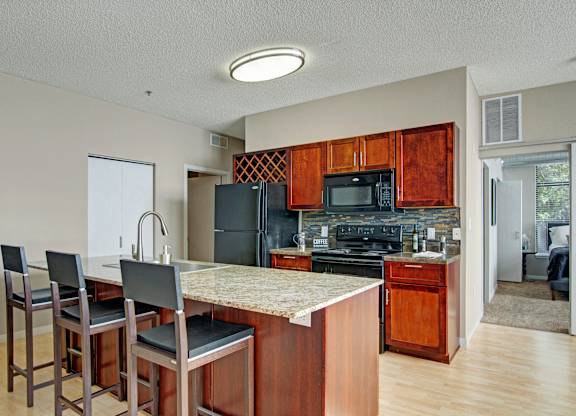 A warm open kitchen here at Marbella on Dean featuring rich wood cabinetry, a spacious island with granite-style countertops and bar seating, a tile backsplash, and a full appliance suite, with light wood flooring, a built-in wine rack, and an adjacent hallway leading toward the bedroom area creating a functional and inviting layout.
