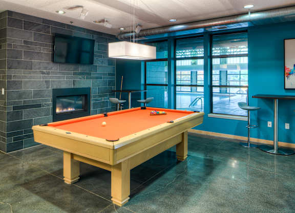 A stylish community game room here at Marbella on Dean featuring a full-size pool table with vibrant felt centered on polished concrete floors, surrounded by teal accent walls, a modern stone fireplace with mounted TV, pendant lighting, and large interior windows that look into the indoor pool area for an energetic and social atmosphere.