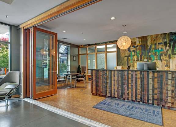 A welcoming leasing office here at Marbella on Dean showcasing a modern wood-paneled front desk, warm pendant lighting, and polished concrete floors, with large glass doors opening to a cozy seating area, contemporary artwork, and natural light that creates an inviting first impression for residents and guests.