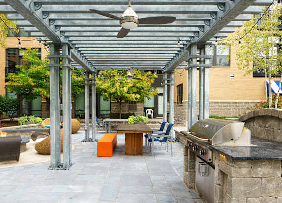 A landscaped outdoor courtyard here at Marbella on Dean showcasing a covered pergola with metal beams and ceiling fans, string lighting, and paved stone flooring, centered around a communal dining table with seating, a built-in grill station, and a cozy fire pit area, all surrounded by mature trees, greenery, and the surrounding apartment buildings for a relaxed gathering space.