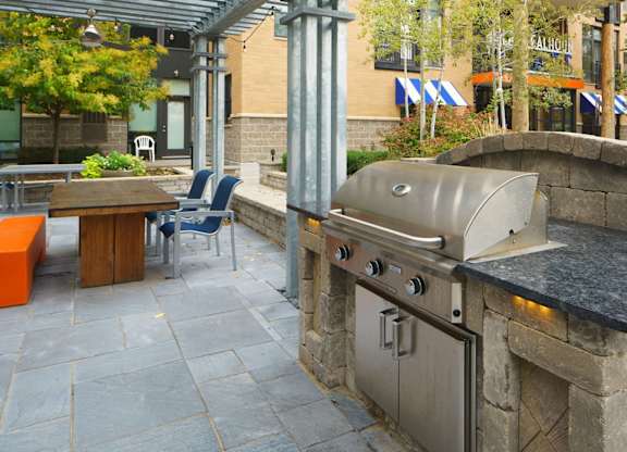 A modern outdoor grilling area here at Marbella on Dean featuring a built-in stainless steel grill set into stone counters, positioned beneath a covered pergola with metal beams and string lighting, alongside stone paver flooring, a wooden communal table with seating, an orange bench, raised planters, and landscaped greenery framed by surrounding apartment buildings.