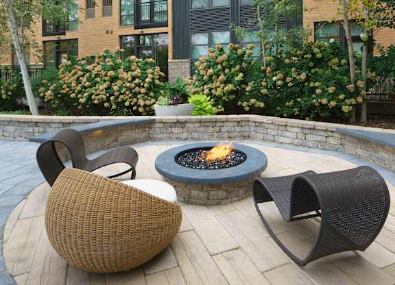 A cozy outdoor fire pit lounge here at Marbella on Dean featuring a circular stone fire pit with a lively flame, surrounded by curved stone seating and sculptural woven lounge chairs set on light stone pavers, with lush landscaped greenery, flowering shrubs, and the surrounding apartment building creating a private and inviting courtyard atmosphere.