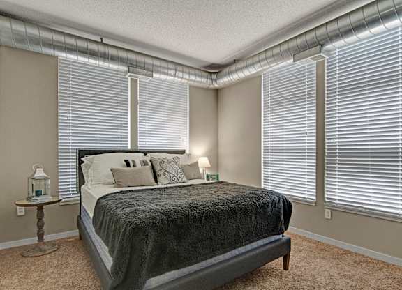 A cozy sunlit bedroom here at Marbella on Dean featuring a plush bed with layered neutral bedding, soft carpeted flooring, and expansive corner windows with blinds that fill the space with natural light, complemented by exposed ductwork along the ceiling, light-toned walls, and a warm, inviting layout ideal for rest and relaxation.
