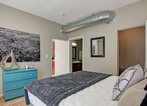 A spacious modern bedroom here at Marbella on Dean featuring a plush bed with layered neutral bedding and accent pillows, soft carpeted flooring, and light-toned walls, highlighted by exposed ceiling ductwork, a bold wall art piece above a teal dresser, and convenient access to a private bathroom and walk-in closet for added comfort and functionality.