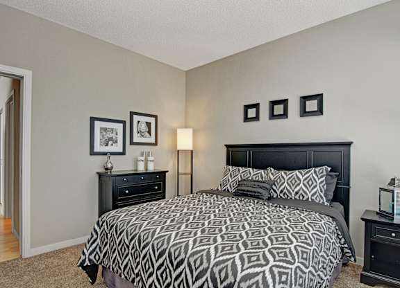 A cozy modern bedroom here at Marbella on Dean featuring a stylish black bed frame with patterned bedding and layered accent pillows, soft carpeted flooring, and neutral-toned walls, complemented by matching nightstands, framed wall art, and a warm floor lamp, with an open doorway leading to the hallway for a comfortable and functional layout.