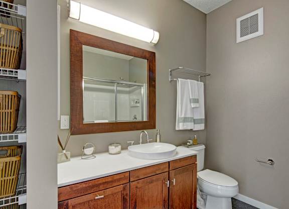 A modern bathroom here at Marbella on Dean featuring a white vanity with warm wood cabinetry, a round vessel sink with sleek fixtures, a large framed mirror with overhead lighting, open shelving with woven baskets, a towel rack, and a glass-enclosed shower creating a clean and functional space.