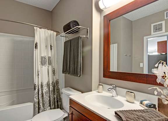 A contemporary bathroom here at Marbella on Dean featuring a white vanity with warm wood cabinetry, an integrated sink with chrome fixtures, a large framed mirror with overhead lighting, neatly folded towels on a wall-mounted rack, a shower with a patterned curtain, and soft neutral walls that create a clean, comfortable atmosphere.