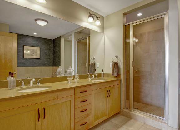 A modern bathroom here at The Mist featuring a wide double-sink vanity with smooth countertops, light wood cabinetry, and ample storage drawers. A large wall mirror and overhead lighting brighten the space, while a glass-enclosed shower and neutral tile flooring create a clean, spa-inspired feel.