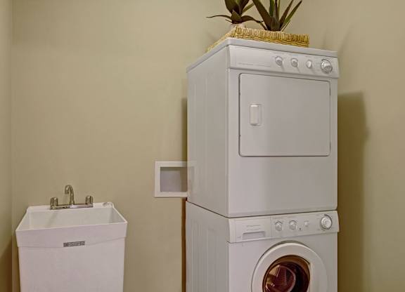 A convenient laundry area here at The Mist featuring a stacked washer and dryer set positioned beside a deep utility sink with a chrome faucet. Neutral wall tones create a clean look, while the compact layout maximizes functionality and provides an efficient space for everyday laundry tasks within the apartment.