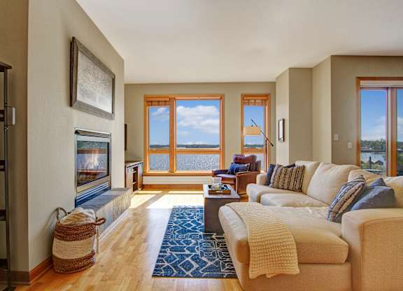 A bright living room here at The Mist featuring light wood-style flooring, a comfortable sectional sofa with textured accent pillows, and a patterned area rug centered around a sleek coffee table. A built-in fireplace adds warmth along one wall, while large windows frame scenic water views and fill the space with natural light.