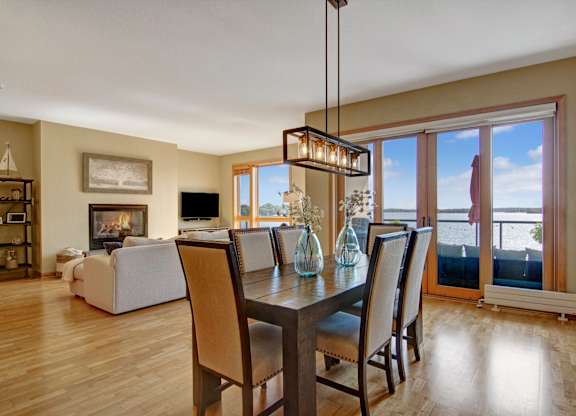A bright open dining and living area here at The Mist featuring light wood-style flooring, a rectangular dining table with upholstered chairs, and a modern pendant light overhead. The space flows into a cozy living room with a fireplace and seating, while large glass doors and windows showcase scenic water views and lead to a private balcony.