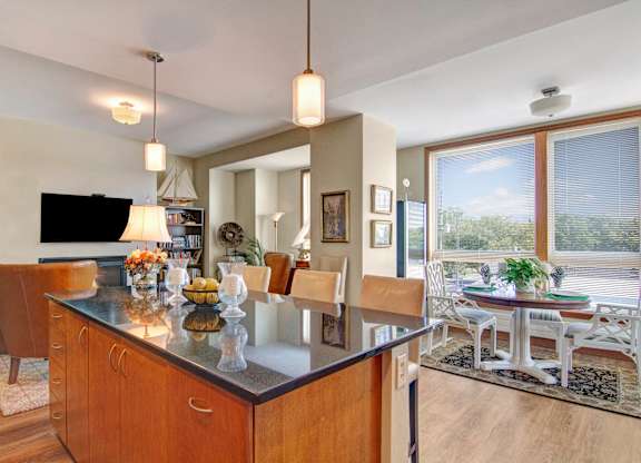 A spacious open kitchen and dining area here at The Mist featuring a large island with bar seating, smooth countertops, and warm wood cabinetry beneath modern pendant lighting. The layout opens to a dining nook with a round table near wide windows, allowing natural light to brighten the space and create an inviting setting for meals and gatherings.