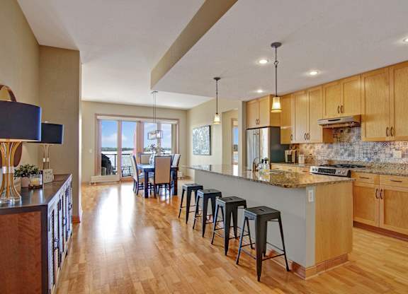 A spacious open kitchen here at The Mist showcasing a long breakfast bar with seating, smooth countertops, and warm wood cabinetry accented by a patterned backsplash. Pendant and recessed lighting brighten the space, while hardwood-style flooring flows into a dining area with large windows and a glass door that bring in abundant natural light.
