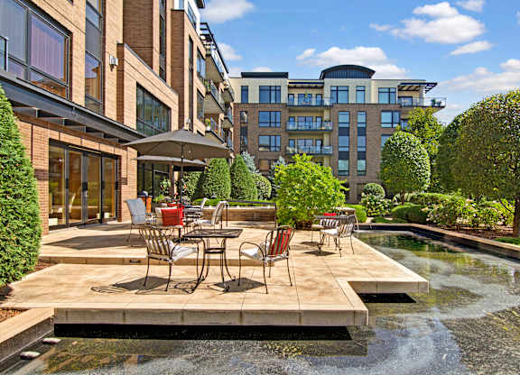 A scenic outdoor patio here at The Mist featuring café-style tables and chairs arranged on a spacious stone terrace overlooking a tranquil water feature. Lush landscaping, manicured trees, and surrounding apartment buildings frame the setting, creating a peaceful space for relaxing outdoors while enjoying fresh air and sunny views.