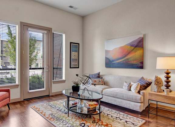 A stylish living room here at Residence at the COR featuring wood-style flooring, a light neutral sofa with patterned accent pillows, a glass coffee table on a colorful area rug, and modern wall art, with large windows and a glass door bringing in natural light and creating a bright, inviting space for relaxing or entertaining.
