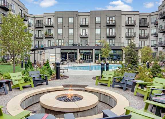 A vibrant outdoor courtyard here at Residence at the COR showcasing a circular fire pit with surrounding seating, landscaped walkways, and colorful lounge chairs, with a resort-style pool and modern apartment buildings with balconies in the background, creating a welcoming community space designed for gathering, relaxation, and outdoor enjoyment.