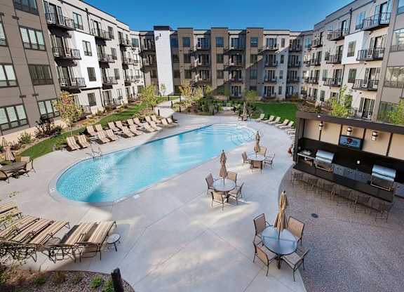 A resort-style pool courtyard here at Residence at the COR featuring a large curved swimming pool with surrounding lounge chairs, shaded patio tables with umbrellas, and a modern outdoor grilling pavilion, all enclosed by contemporary apartment buildings with balconies and landscaped green spaces that create an inviting setting for relaxation and socializing.