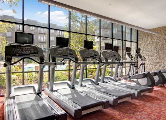 A sunlit fitness center here at Residence at the COR featuring a row of modern treadmills and cardio machines positioned along expansive floor-to-ceiling windows, with views of the landscaped courtyard and pool area, complemented by textured stone accent walls and an open layout that creates an energizing space for daily workouts.