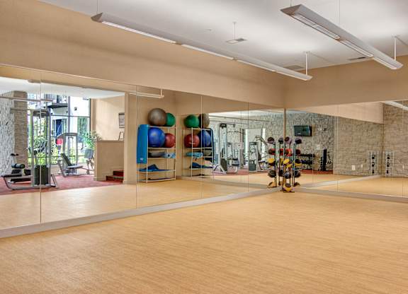 A spacious fitness studio here at Residence at the COR featuring a wide open workout area with smooth flooring, full mirrored walls, and overhead lighting, complemented by neatly organized exercise balls, mats, and free weights, with adjacent cardio and strength equipment visible through the mirrors, creating a versatile space ideal for stretching, group classes, and functional training.