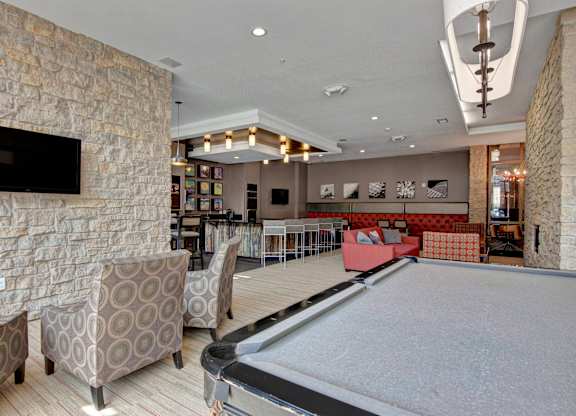 A stylish resident lounge here at Residence at the COR featuring a spacious open layout with a pool table, patterned lounge chairs, and a mounted television, complemented by stone accent walls, warm wood finishes, a modern bar with seating, cozy sofas, and contemporary lighting that creates an inviting space for relaxing, socializing, and entertainment.