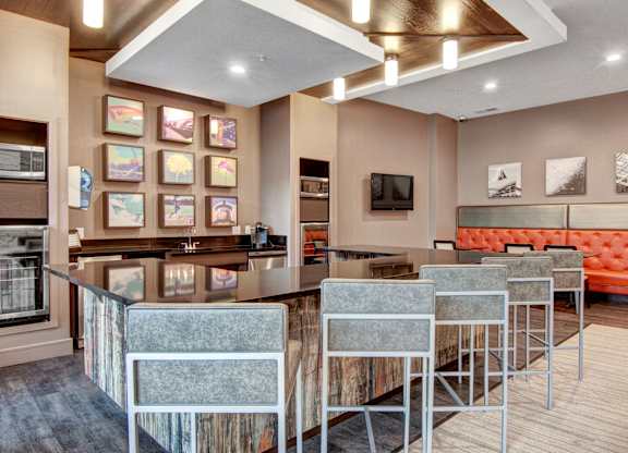 A contemporary resident lounge here at Residence at the COR showcasing a bar-height island with cushioned stools and sleek countertops, complemented by warm wood accents, an orange tufted banquette, modern wall art, and layered recessed and pendant lighting, all within an open, inviting layout designed for socializing, entertaining, and everyday relaxation.