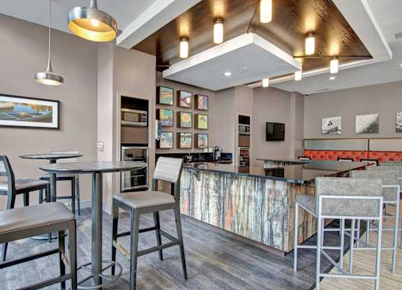 A stylish resident lounge here at Residence at the COR featuring a bar-height island with sleek countertops and modern stools, complemented by café-style tables, warm wood ceiling accents, contemporary pendant lighting, colorful framed artwork, and an inviting seating area that creates a polished, social setting for gathering, entertaining, or unwinding.