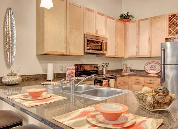 A contemporary kitchen here at Residence at the COR showcasing light wood cabinetry, sleek countertops with an integrated double sink, stainless-style appliances, and a built-in microwave, complemented by a breakfast bar set with place settings, decorative accents, and warm pendant lighting that creates a functional and inviting cooking space.