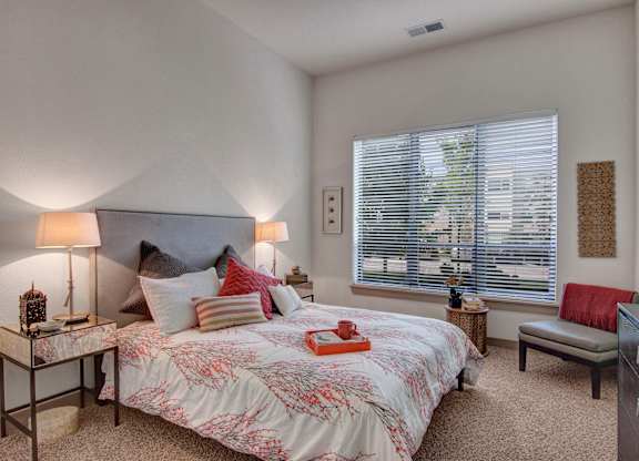 A serene bedroom here at Residence at the COR featuring a plush bed with layered neutral and accent pillows, a patterned comforter, and matching bedside tables with soft-glow lamps, complemented by cozy carpet, a large window with blinds letting in natural light, and thoughtfully placed seating and decor that create a calm, inviting retreat.