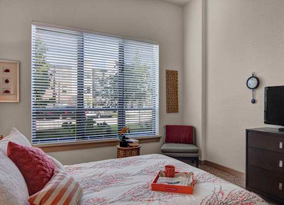A comfortable bedroom here at Residence at the COR featuring a neatly made bed with layered neutral and warm accent pillows, a patterned comforter, and a small tray accent, complemented by plush carpeting, a large window with blinds bringing in natural light, a cozy accent chair, and a dresser with a mounted television creating a functional and inviting retreat.