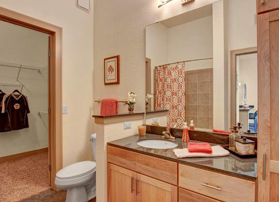 A clean bathroom here at Residence at the COR featuring a wood-tone vanity with a smooth countertop and single sink, a large wall mirror with overhead lighting, neatly arranged bath accessories and towels, and a shower with tiled surround and patterned curtain, with an adjacent walk-in closet visible that adds convenience and functional storage.