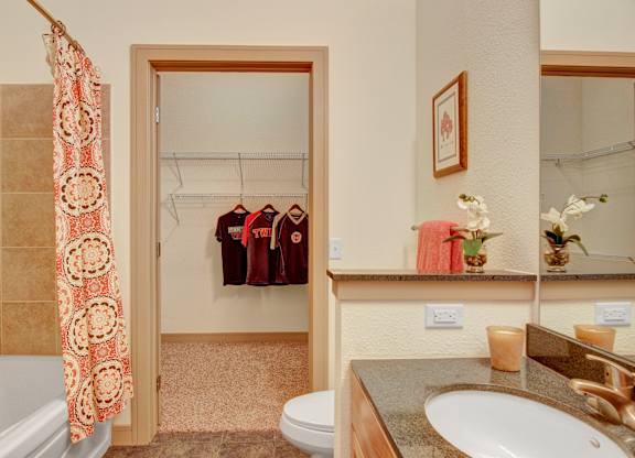 A bright bathroom here at Residence at the COR showcasing a wood-tone vanity with a smooth countertop and single sink, a wide wall mirror, and a tub with tiled surround and patterned shower curtain, while an open doorway leads to a spacious walk-in closet with shelving and hanging space for added storage and everyday convenience.