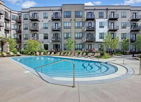 A resort-style outdoor pool here at Residence at the COR featuring a curved entry with handrails and shallow steps, surrounded by a wide concrete deck lined with lounge chairs, while a modern apartment building with private balconies and landscaped greenery creates a polished, inviting backdrop for relaxation and social time.