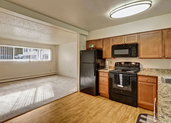 Cozy apartment interior here at Montclair Apartments with an open layout. The kitchen has wooden cabinets, black appliances, and a granite countertop. Large window with blinds. Bright and inviting.