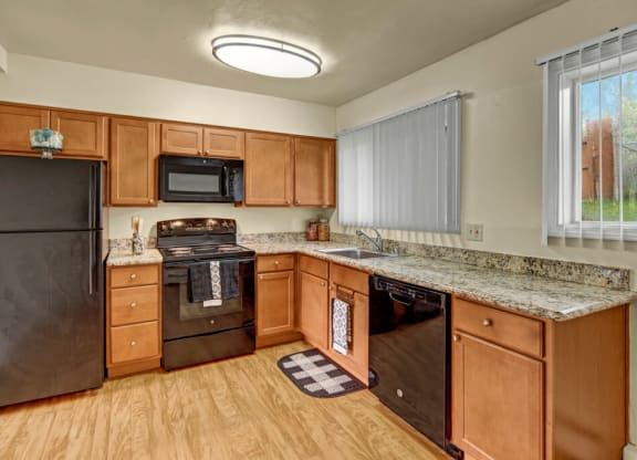 A modern kitchen here at Monctlair Apartments with wooden cabinets, black appliances including a fridge, stove, and dishwasher, granite countertops, and a window with vertical blinds.