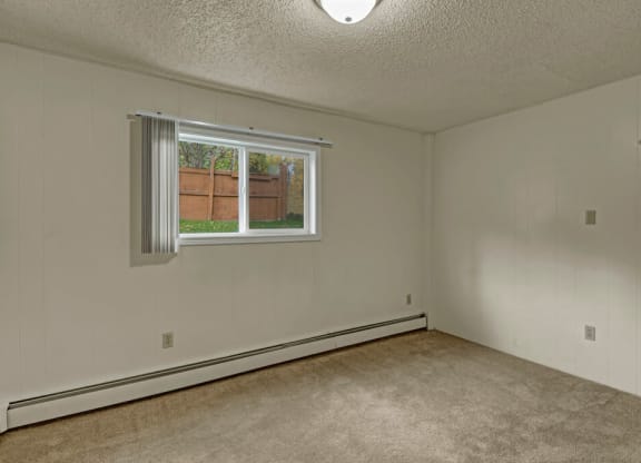 Empty bedroom here at Montclair Apartments with beige carpet and off-white walls, featuring a single window with vertical blinds and a view of a wooden fence and greenery outside. Neutral and calm atmosphere.
