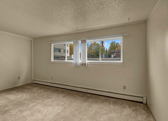 Empty bedroom here at Montclair Apartments with light beige walls and carpet, large window with vertical blinds showing trees and a building outside. Calm, neutral atmosphere.