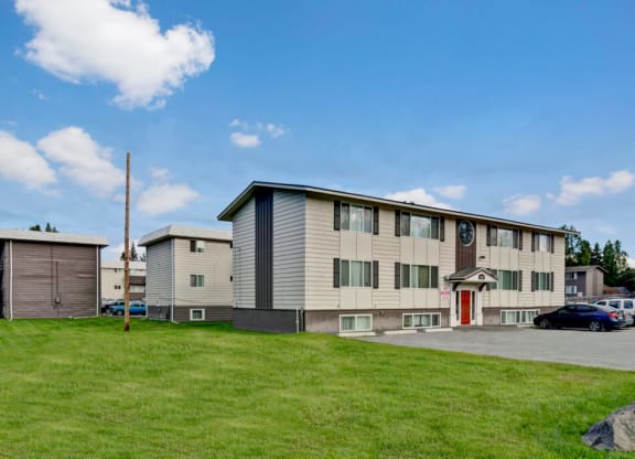 Two-story Montclair apartment building with white siding and red door set against a blue sky with fluffy clouds. Surrounding green lawn adds a serene touch.
